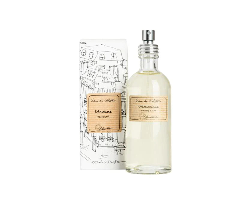 A clear glass bottle of Lothantique Verbena eau de toilette by Lothantique next to its illustrated packaging, displaying sketches of buildings and cursive text. The liquid in the bottle is pale.
