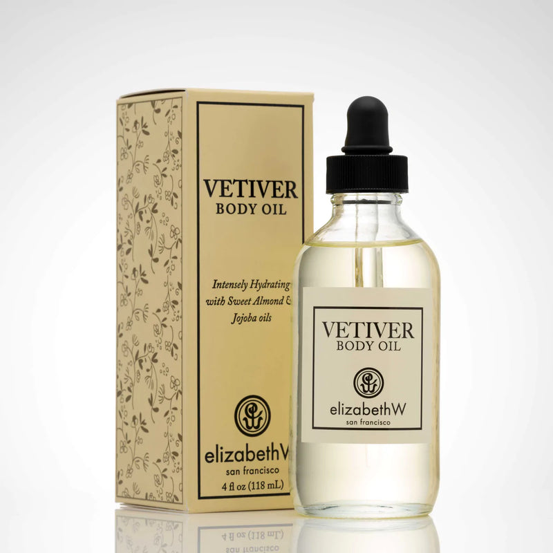 A clear glass bottle of elizabeth W Signature Vetiver Body Oil with a dropper, next to its decorative beige and gold box, both labeled "elizabeth W San Francisco." The box features intricate floral designs.