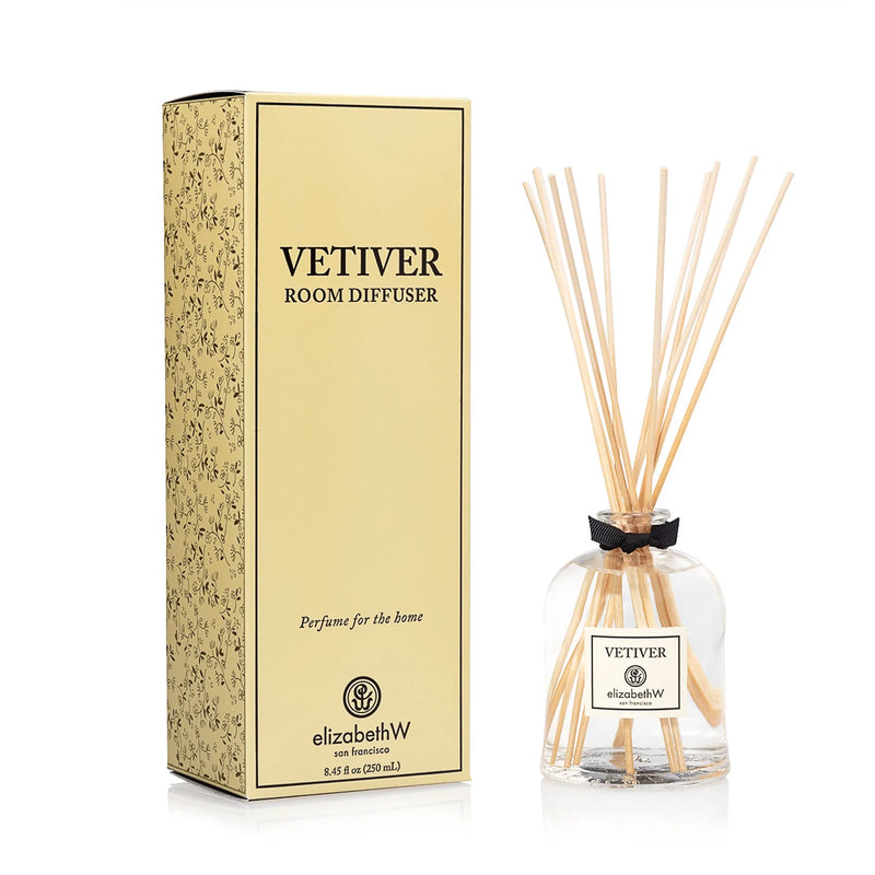 A elizabeth W Signature Vetiver Diffuser by elizabeth W next to its packaging. The environmentally friendly diffuser consists of a clear glass bottle with a gold label and multiple wooden sticks inserted inside. The tall,