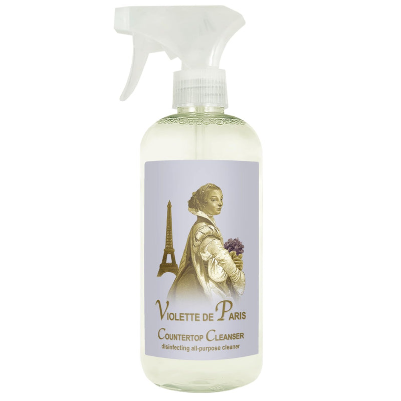 A transparent spray bottle labeled "La Bouquetiere Violette de Paris CounterTop Cleanser" featuring an illustration of a vintage woman, the Eiffel Tower, and essential oils.