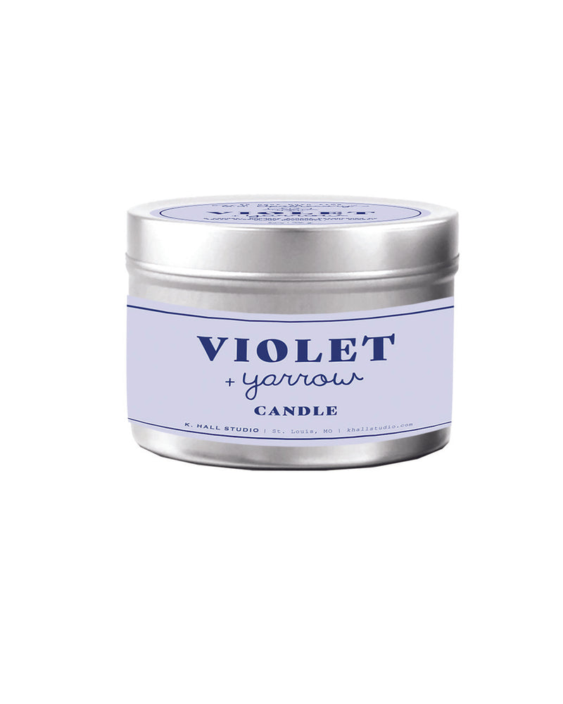 A cylindrical floral candle in a silver tin with a pale purple label reading "U.S. Apothecary Violet + Yarrow Tin Candle" against a white background.