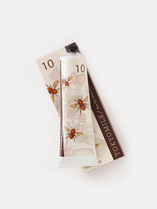 A tube of TokyoMilk Honey and The Moon No. 10 Bon Bon Shea Butter Lotion in floral packaging, featuring illustrations of delicate pink flowers and bees, set against a white background, with hints of sugared violet in the design by Margot Elena