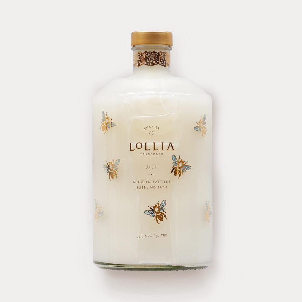 A bottle of Margot Elena's Lollia Wish Bubble Bath with a cork stopper, featuring a clear design adorned with golden bee and butterfly illustrations. The label reads “Sugared Pastille” and includes Vanilla Bean.