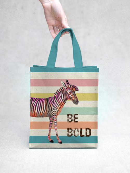 A hand holding a TokyoMilk Zebra Be Bold Small Tote with a colorful striped design featuring a zebra with pink and black stripes next to the phrase "be bold" by Margot Elena.